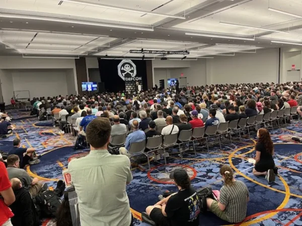 Image of a crowded room of attendees with a large DefCon banner at the front of the room.